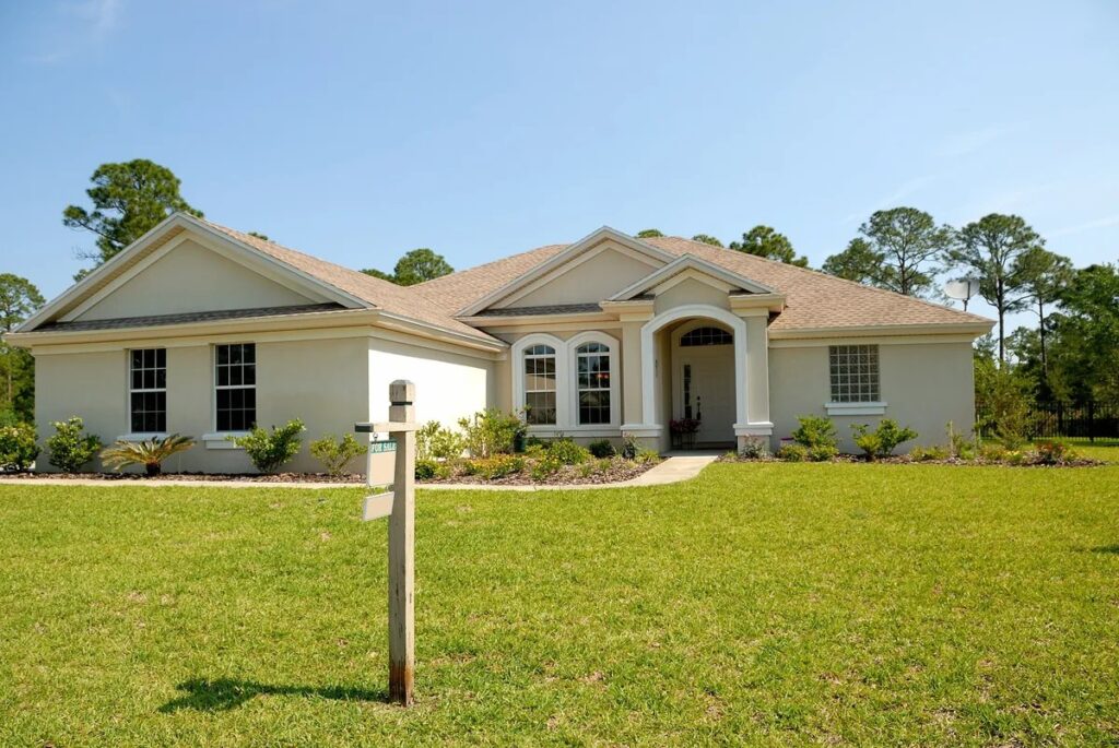 Single-story beige house with hip roof, arched entrance, green lawn, and wooden sign post under a blue sky.
