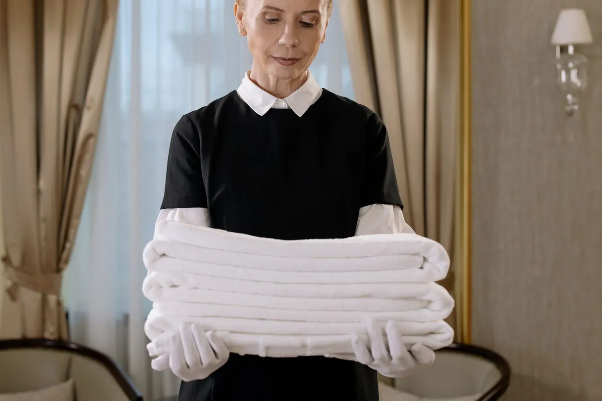 Housekeeper in dark uniform and white gloves holding folded white towels in a hotel-style room.