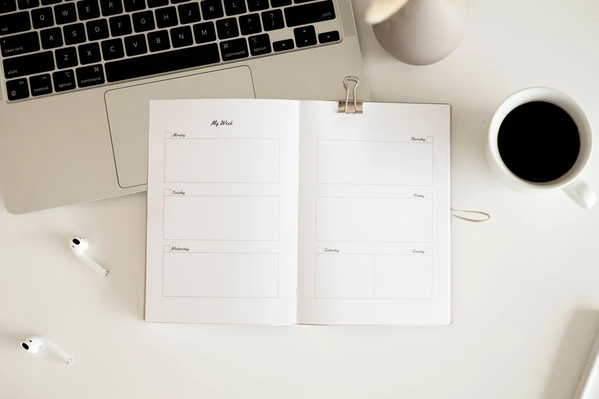 Open weekly planner with blank day boxes on a white desk beside a laptop keyboard, coffee, and earbuds.