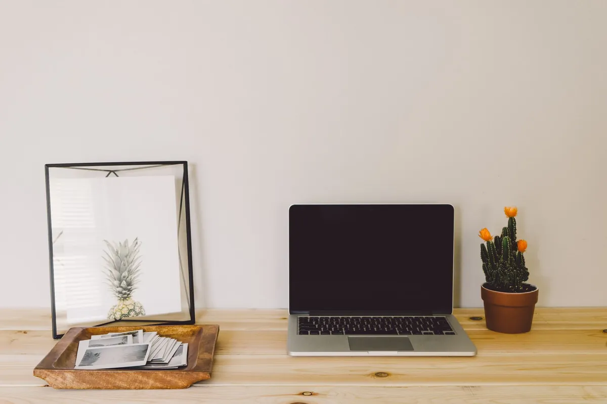 A laptop on a wooden desk with papers, a small cactus, and a framed pineapple print.